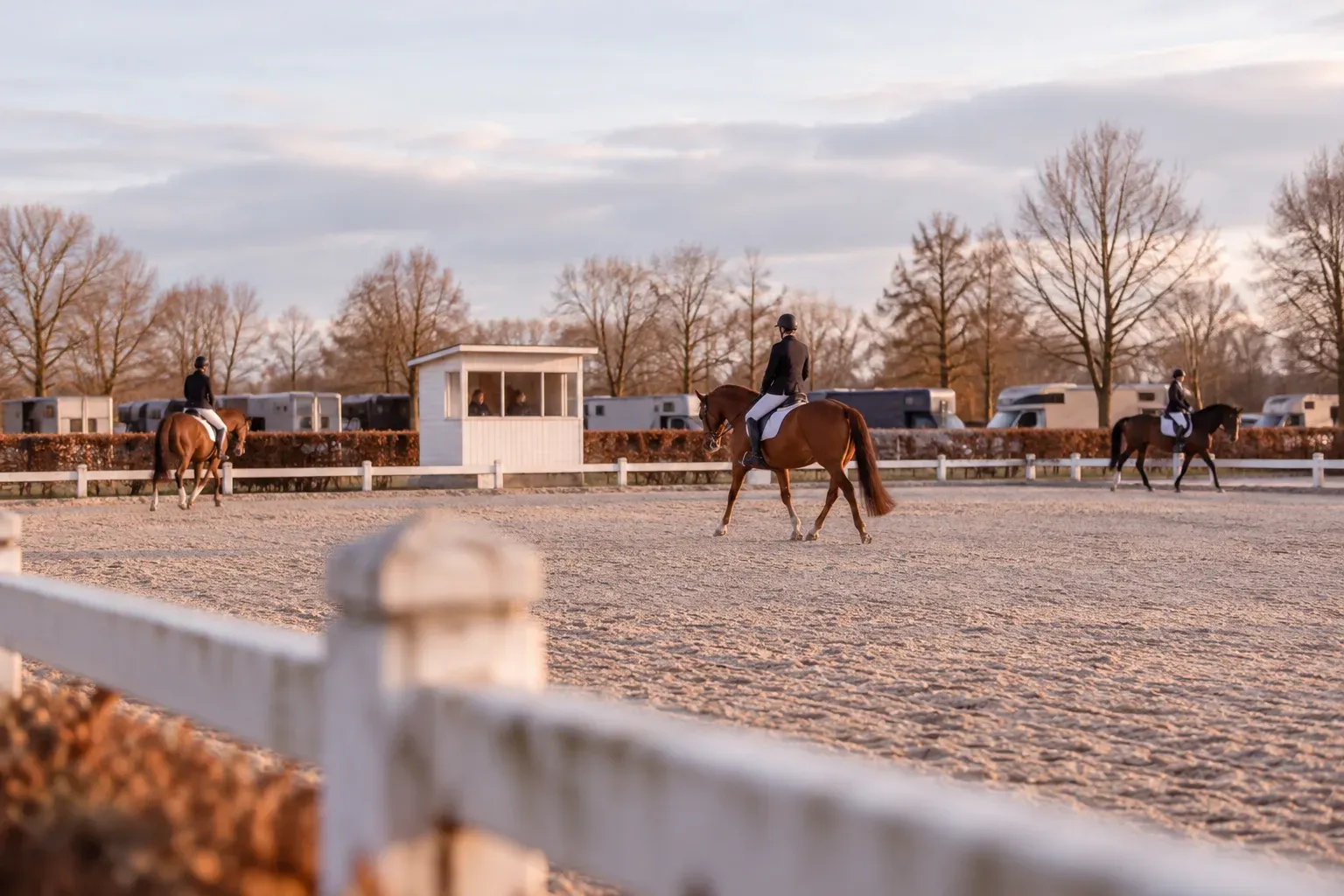 Ruiters rijden los op een wedstrijdterrein, met het juryhuisje en paardentrailers op de achtergrond
